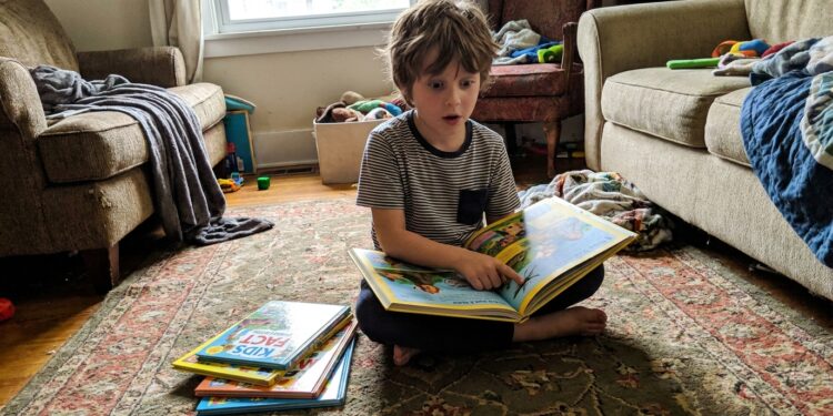Boy sitting on living room floor reading a colorful illustrated fact book, surprised, with a stack of kids trivia books beside him